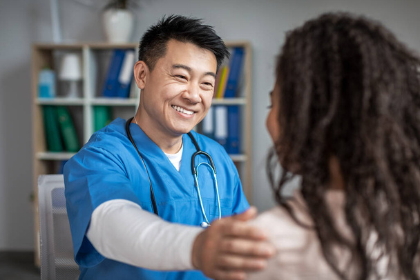 Smiling adult chinese male doctor calms and supports young african american woman patient in clinic office interior. Great service, examination with therapist, treatment of ill, medical health care - Zdjęcie, obraz