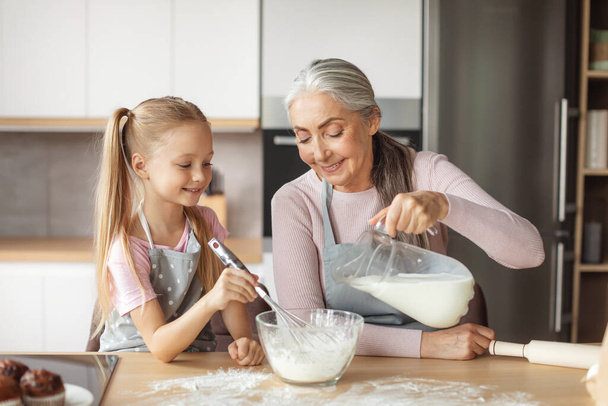 Happy european elderly grandmother and little granddaughter in aprons pour milk to dough in kitchen interior. Homemade sweet food, cooking lesson together for family at home, hobby and housekeeping - Fotó, kép