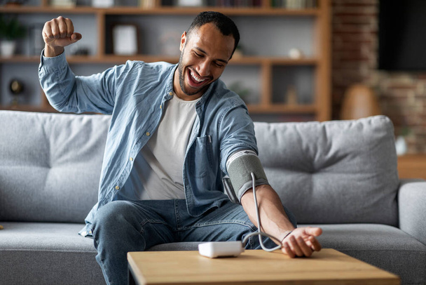 Excited Young Black Man Checking Blood Pressure With Electronic Monitor At Home, Happy African American Guy Using Sphygmomanometer While Sitting On Couch In Living Room, Feeling Good And Healthy - 写真・画像