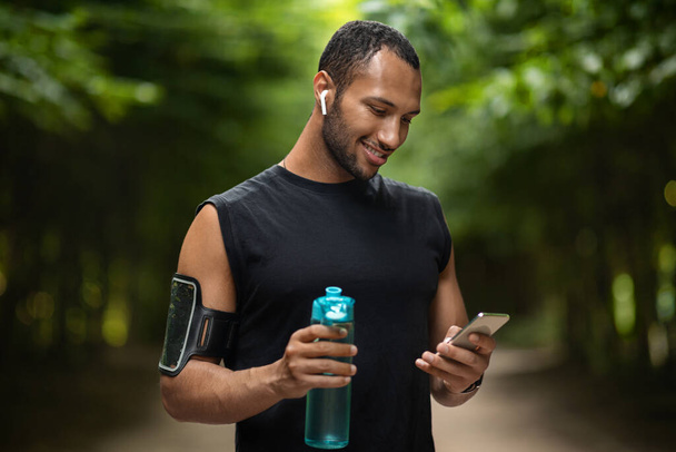 Smiling young black sportsman exercising at public park, using smartphone and wireless earbuds, checking mobile fitness application, drinking water while having break, copy space - Foto, Imagem