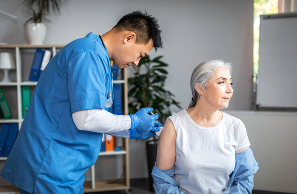 Серйозний дорослий азіатський лікар робить ін'єкції старій жінці пацієнта в інтер'єрі клінічного кабінету. Вакцинація та імунізація пенсіонерів, лікування, медична допомога під час пандемії грипу та ковадла-19
 - Фото, зображення