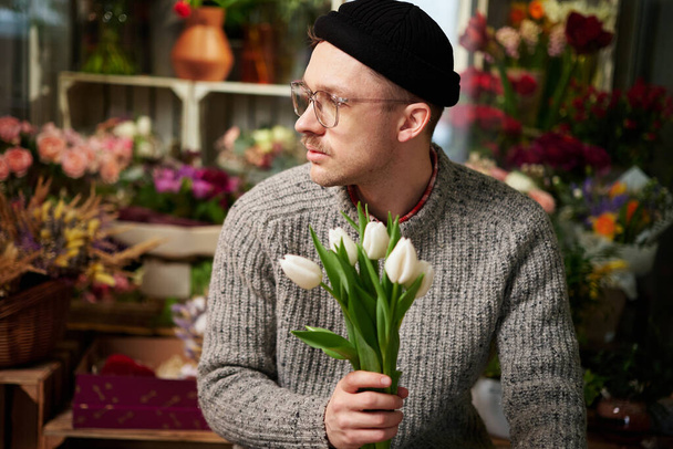 Bearded caucasian hipster male florist in sweater holding beautiful bouquet of spring white tulip bouquet. Valentines Day, love, romantic or International Womens Day concept. High quality image - Foto, Bild