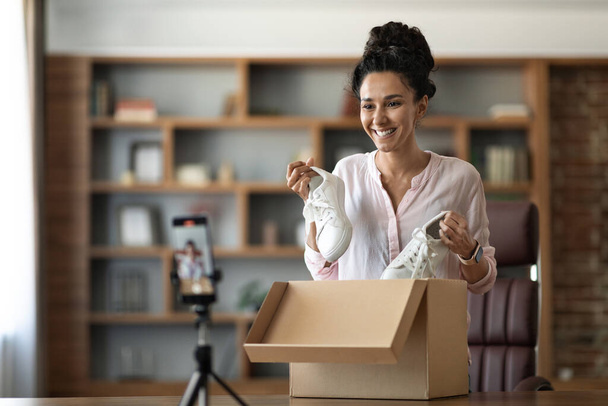 Cheerful pretty young woman famous vlogger streaming from home while unpacking parcel, happy brunette lady holding shoes and recording video on smartphone, copy space. Social media and business - Photo, image