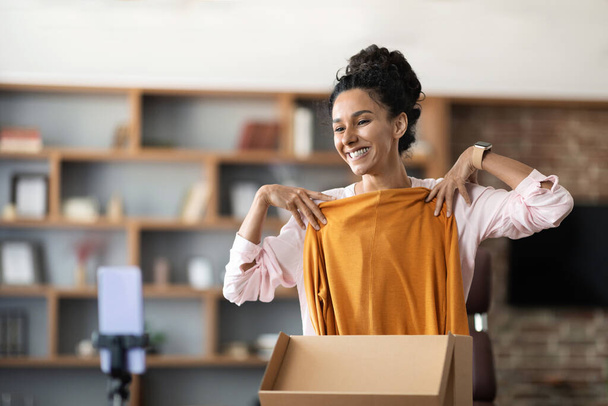 Happy attractive young woman unpacking delivery box and recording video on smartphone set on tripod, excited lady having video chat with friend, showing her new shirt and smiling, copy space - Φωτογραφία, εικόνα