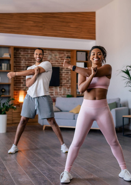 Smiling young african american male and female in sportswear doing stretching or warm up exercises for hands in living room interior. Body care together, fitness at home weight loss during covid-19 - Фото, изображение