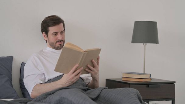 Yatakta otururken sakallı genç adam kitap okuyor. - Fotoğraf, Görsel