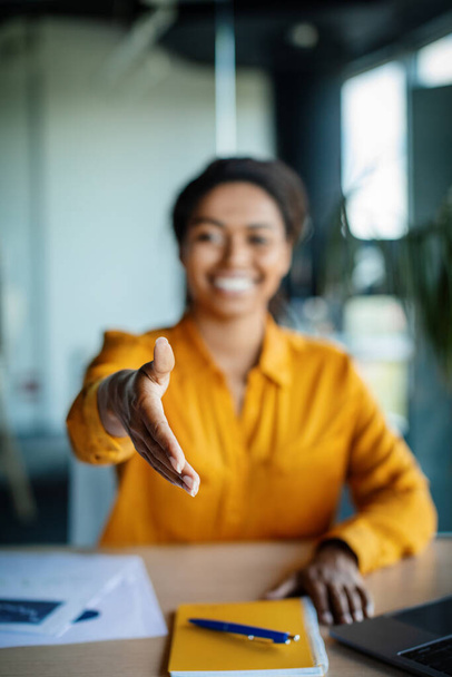 Cooperation concept. Happy black businesswoman extending hand for handshake at camera, sitting at desk in office. Female entrepreneur offering partnership, greeting companion - Photo, Image
