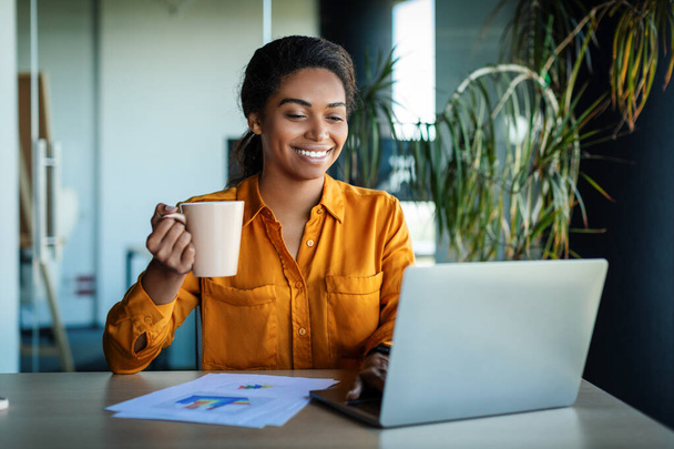 Happy african american businesswoman working on laptop and drinking coffee, sitting at workplace in office. Technology and business career concept - Foto, immagini