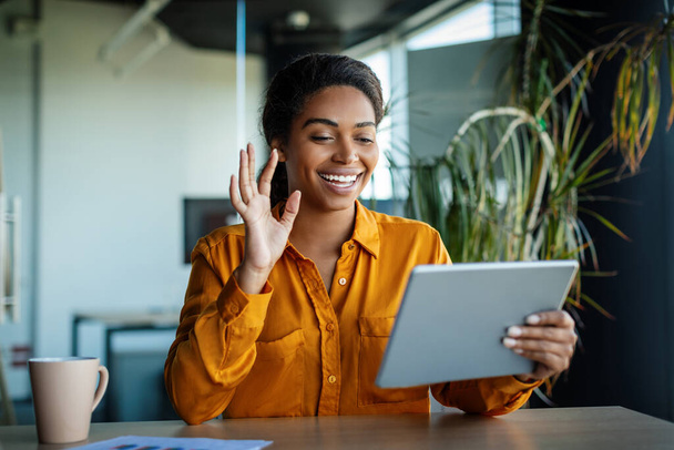 Happy black businesswoman sitting at table in office, having video chat with business partners, using digital tablet, smiling at gadget screen and gesturing - Foto, imagen