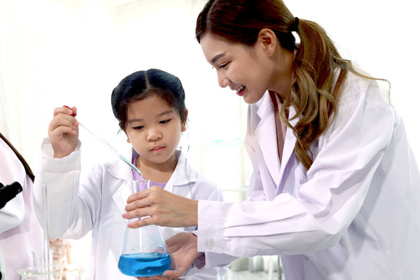 Student and teacher in lab coat have fun together while learn science experiment in laboratory. Young adorable Asian scientist kid adding some chemical to blue flask with pipette, little children students study research at school lab. Science educati - Foto, immagini