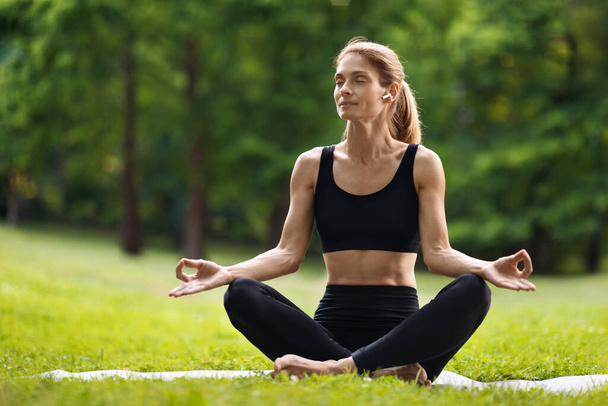 Peaceful blonde woman in black tight sportswear sitting on fitness mat with closed eyes in lotus pose, practicing yoga at public park, listening to music, using brand new earbuds, copy space - Foto, Bild