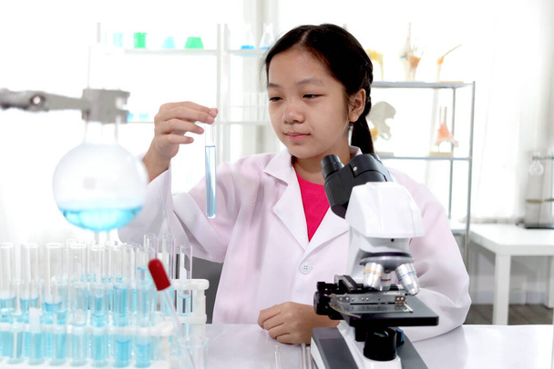 Adorable schoolgirl in lab coat doing science experiments, young scientist looking at blue test tube and learning science experiment in laboratory. Asian kid using microscope, apparatus and lab equipment for research. Science education for kid concep - Фото, зображення