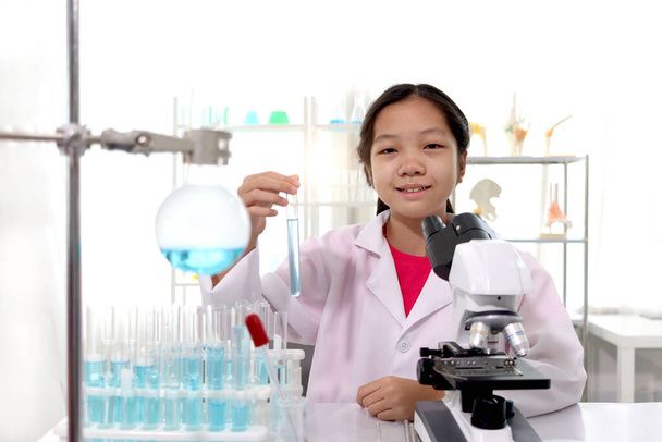 Adorable schoolgirl in lab coat doing science experiments, young scientist showing at blue test tube and learning science experiment in laboratory. Asian kid using microscope, apparatus and lab equipment for research. Science education for kid concep - Foto, Bild
