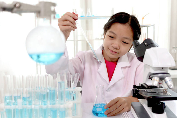 Adorable schoolgirl in lab coat doing science experiments, young scientist adding some chemical to blue flask with pipette and learning science experiment in laboratory. Asian kid using microscope, apparatus and lab equipment for research. Science ed - Foto, Imagen
