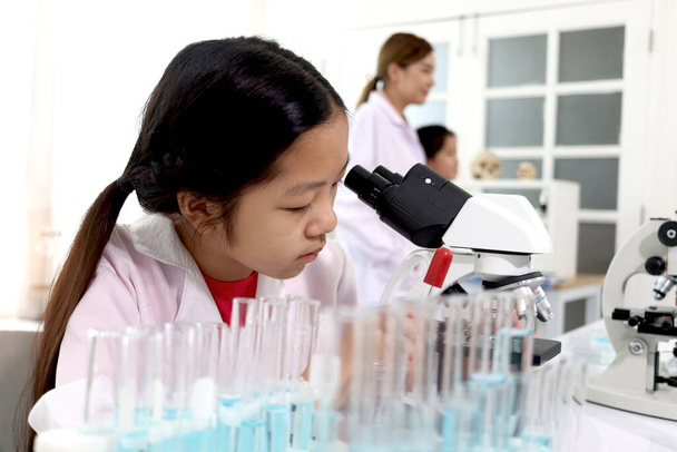 Adorable schoolgirl in lab coat doing science experiments, young scientist looking through microscope and learn science experiment in laboratory. Asian kid using apparatus and lab equipment for research with teacher and classmate as blur background.  - Фото, зображення