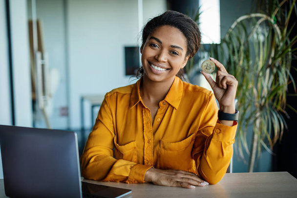 Excited black businesswoman holding golden bitcoin and smiling at camera, sitting at desk with laptop computer, trading on stocks and markets while working at office. Cryptocurrency concept - Valokuva, kuva