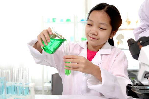 Adorable Asian schoolgirl in lab coat doing science experiments, pouring green reagent solution from beaker into test tube, happy smiling young scientist kid having fun during doing experiments, child using microscope, apparatus and lab equipment for - Photo, Image