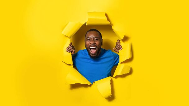 African american man shouting, reacting on something amazing posing through hole in yellow paper background, panorama. Emotions concept - Фото, зображення