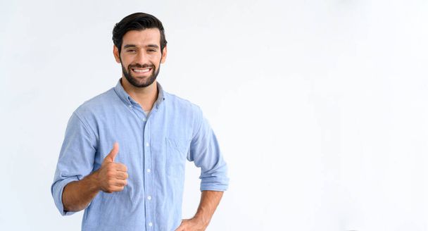 Handsome young bearded man in casual blue t-shirt smiling and showing thumb up sign, looking at the camera, standing isolated on white background - Foto, Imagem