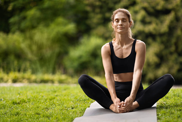 Peaceful slender blonde middle aged woman in black tight sportswear sitting with closed eyes on fitness mat, stretching body outdoors, panorama with copy space. Open air workout concept - Foto, imagen