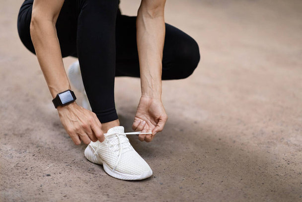 Cropped shot of woman in black sportswear with fitness bracelet tying shoelaces on stylish white sneakers before running outdoors, copy space. Sports shoes concept - 写真・画像
