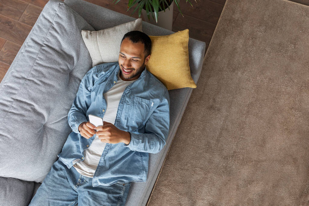 Black Happy Man With Smartphone Relaxing On Comfortable Couch At Home, Top View Of Smiling Young African American Guy Messaging On Mobile Phone While Resting On Sofa In Living Room, Copy Space - Фото, зображення