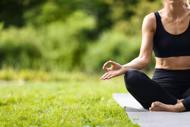 Unrecognizable slim sporty woman in black tight sportswear sitting in lotus pose on fitness mat, meditating outdoors in public park or forest, panorama with copy space. Open air yoga concept - Valokuva, kuva