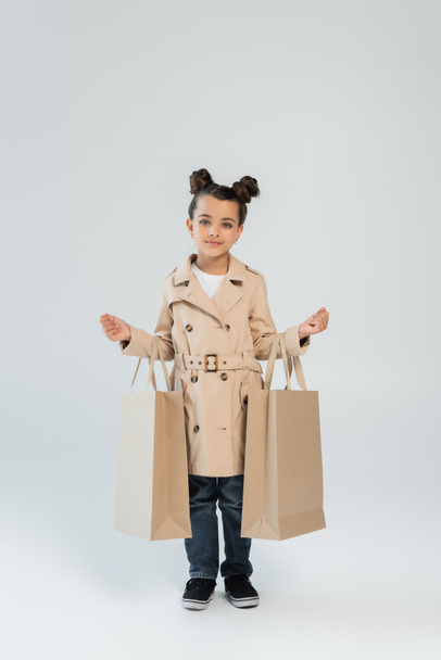 full length of stylish child in trench coat and jeans holding shopping bags on grey, black friday concept - Zdjęcie, obraz