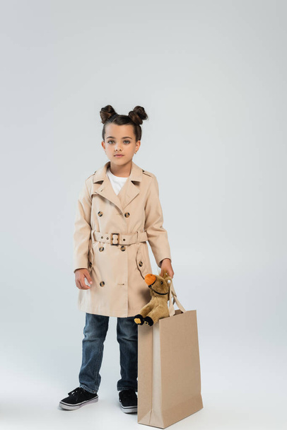 full length of stylish child in trench coat and jeans holding shopping bag with toy horse on grey - Foto, Bild
