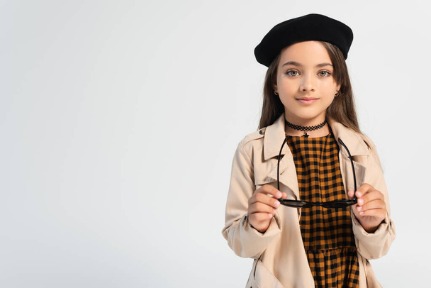 happy girl in stylish trench coat and beret holding sunglasses isolated on grey - Foto, Imagen