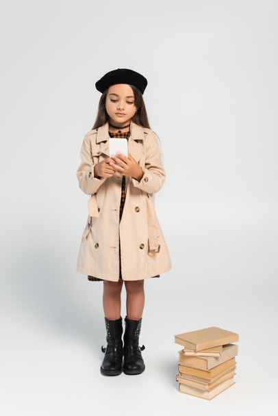 full length of cute kid in stylish autumnal outfit and beret using smartphone near stacked books on grey - Φωτογραφία, εικόνα