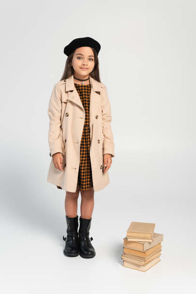full length of smiling kid in stylish autumnal outfit and beret standing near stacked books on grey - Foto, imagen