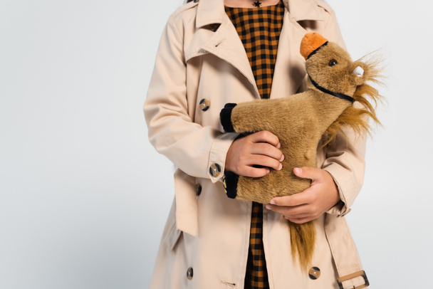 cropped view of girl in stylish trench coat standing with toy horse isolated on grey - Foto, Imagem