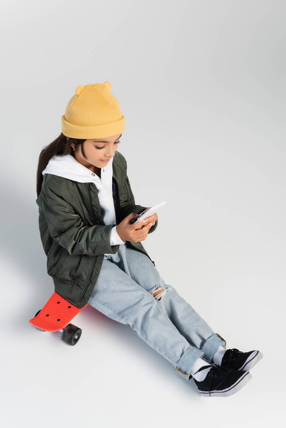 high angle view of cute girl in trendy autumnal outfit using smartphone and sitting on penny board on grey - Valokuva, kuva