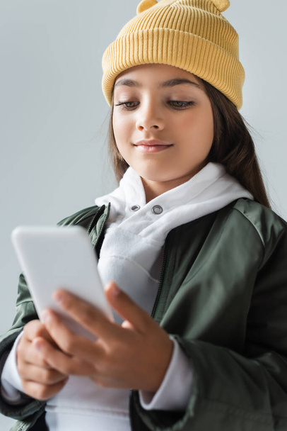 cheerful kid in trendy autumnal outfit and beanie hat using smartphone isolated on grey - Foto, immagini