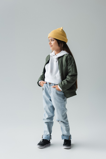 full length of smiling kid in yellow beanie hat and stylish autumnal outfit posing with hands in pockets on grey - Photo, Image