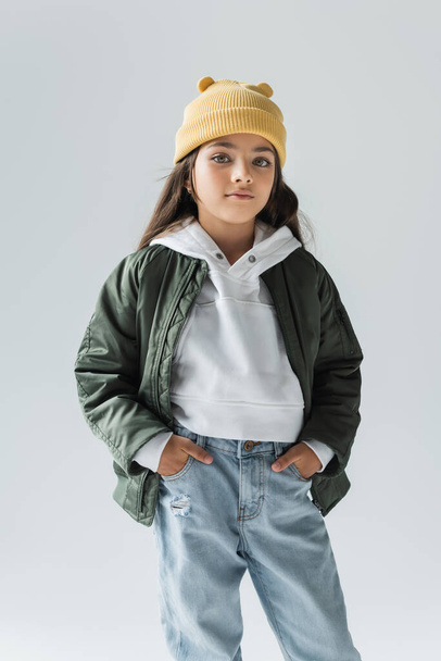 portrait of cute kid in yellow beanie hat and stylish autumnal outfit posing with hands in pockets isolated on grey  - Foto, Bild