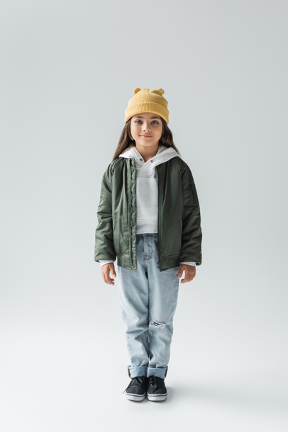 full length of adorable girl in trendy autumnal outfit smiling and standing on grey - Photo, Image