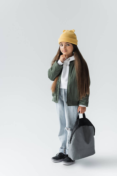 full length of adorable girl in trendy autumnal outfit and beanie hat holding backpack on grey - Фото, изображение