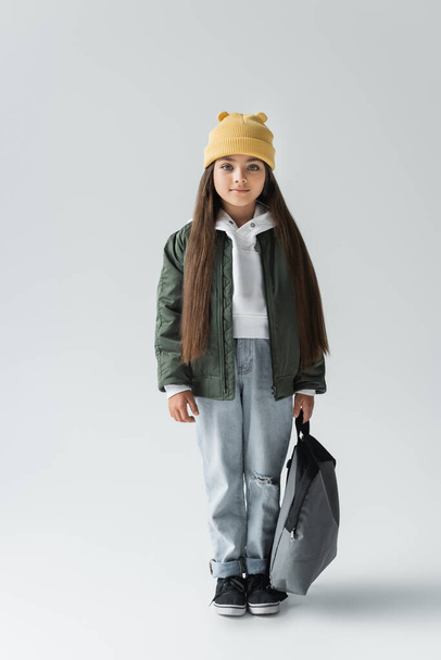 full length of smiling and cute girl in trendy autumnal outfit holding backpack on grey - Foto, immagini