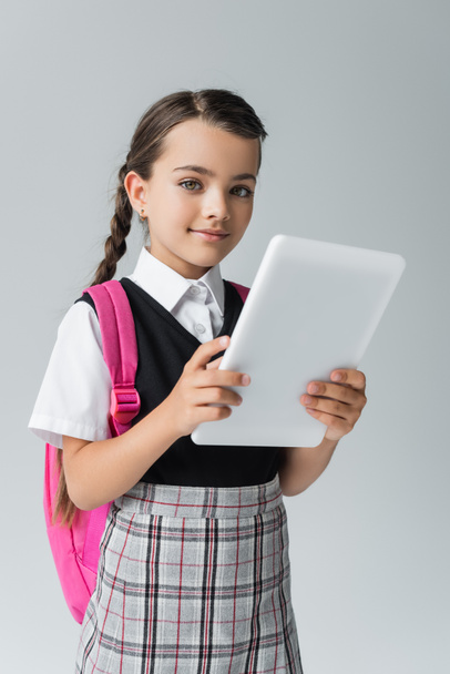 happy and cute schoolgirl in uniform holding digital tablet while smiling isolated on grey - Foto, Bild