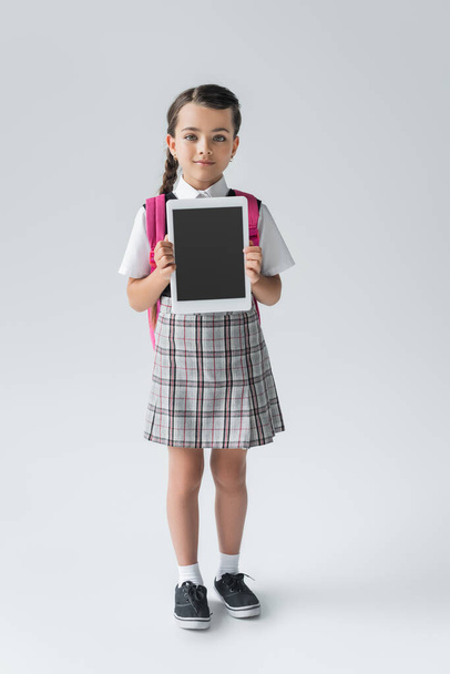 full length of cute schoolgirl in uniform holding digital tablet with blank screen on grey - Photo, image