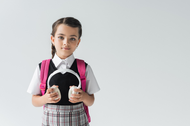 schoolgirl in uniform standing with backpack and holding wireless headphones isolated on grey  - Фото, зображення