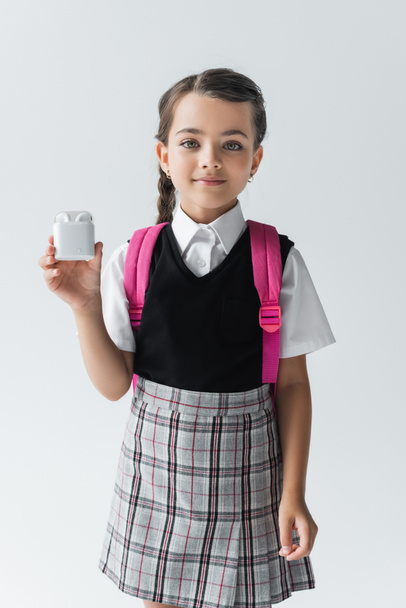 cheerful schoolgirl in uniform standing with backpack and holding earphone case isolated on grey - Valokuva, kuva