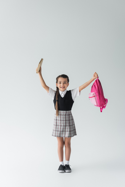 full length of excited schoolgirl in uniform holding pink backpack and book on grey  - Photo, Image