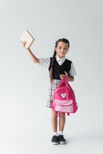 full length of cute schoolgirl in uniform holding pink backpack and book above head on grey  - 写真・画像