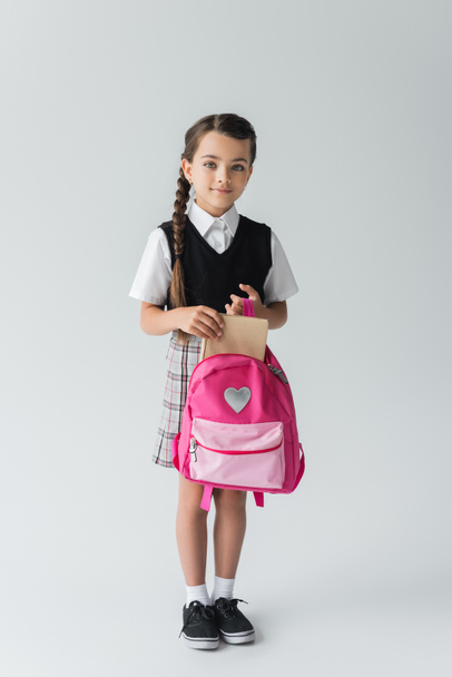 full length of cute schoolgirl in uniform holding pink backpack and book on grey  - Foto, Imagen