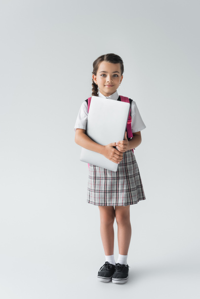 full length of schoolgirl in uniform standing with backpack and laptop on grey - Φωτογραφία, εικόνα