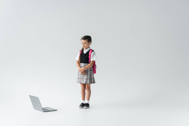 full length of schoolgirl in uniform standing with backpack and looking at laptop on grey - Foto, immagini