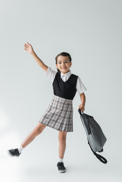 full length of adorable and cheerful schoolgirl in uniform holding backpack on grey - Fotó, kép